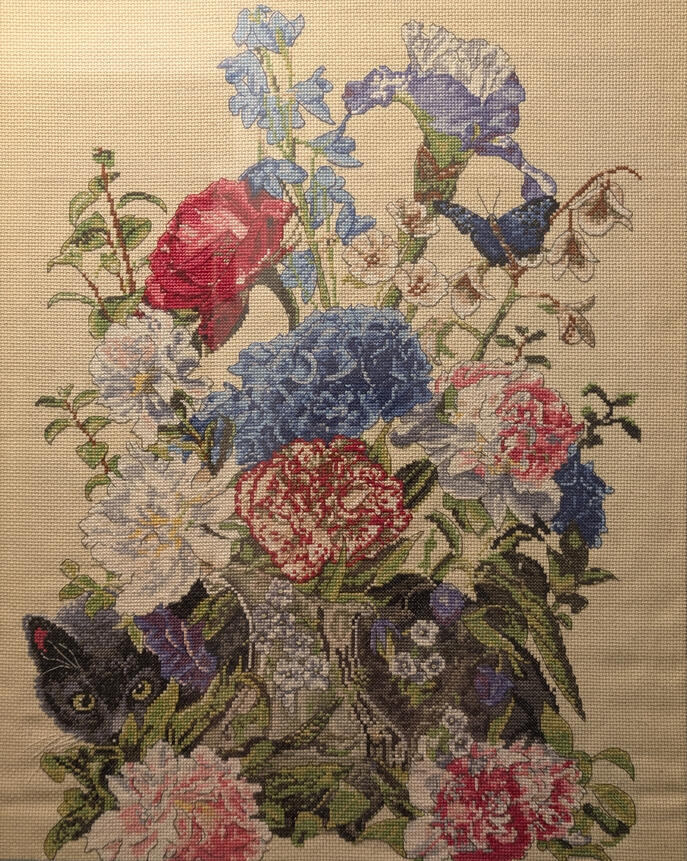 A cross stitch image of a vase with flowers; a cat is hiding behind the vase.